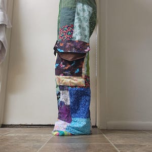 May include: Full-length patchwork pants featuring a variety of fabric squares in shades of green, blue, purple, and yellow. The design includes patterns of leaves, butterflies, and floral motifs, with a drawstring waist and a large brown pocket flap.