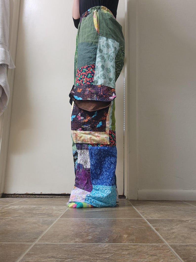 May include: Full-length patchwork pants featuring a variety of fabric squares in shades of green, blue, purple, and yellow. The design includes patterns of leaves, butterflies, and floral motifs, with a drawstring waist and a large brown pocket flap.