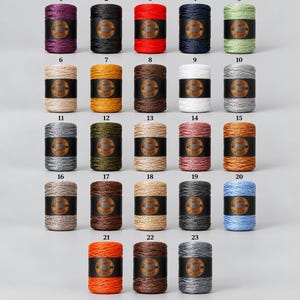 Puede incluir: Veintitrés bobinas de hilo cilíndricas dispuestas en una cuadrícula, cada una envuelta con una etiqueta negra que presenta un logotipo circular. Los colores del hilo incluyen morado, negro, rojo, azul marino, verde claro, beige, amarillo, marrón, blanco, plata, gris, oliva, tostado, rosa, naranja y azul.