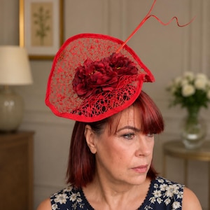 May include: A vibrant red fascinator hat featuring a wide, circular brim with an intricate, web-like mesh pattern. The headpiece is adorned with a cluster of deep red fabric flowers and a single, thin, curved red wire accent extending upwards.