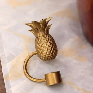 Puede incluir: Un tap&oacute;n de botella dorado con forma de pi&ntilde;a. El tap&oacute;n tiene un cuerpo texturizado y un asa curva que se conecta a una base cil&iacute;ndrica. La corona de la pi&ntilde;a est&aacute; detallada con hojas puntiagudas.