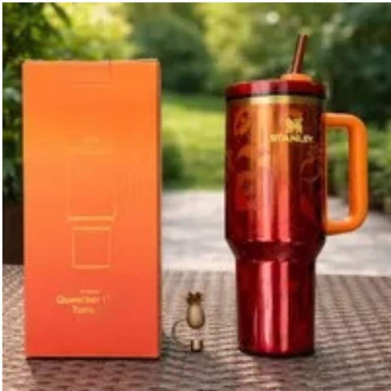 Puede incluir: Un vaso Stanley Quencher rojo y naranja con asa y pajita, junto a su caja naranja. El vaso tiene un acabado met&aacute;lico y el logotipo de Stanley. La caja tiene las palabras "Quencher" y "Tote".