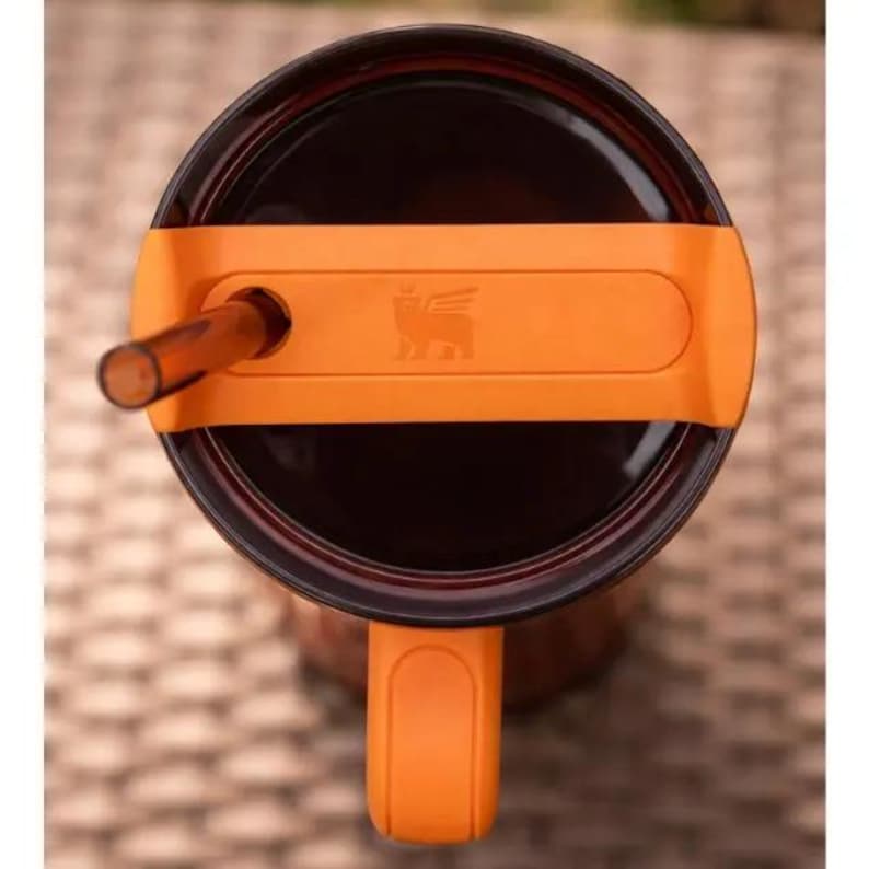 Puede incluir: Vista superior de un termo de viaje Stanley con tapa y asa naranjas. La taza contiene un l&iacute;quido oscuro y una pajita transparente sobresale de la tapa. La tapa tiene el logotipo de Stanley.
