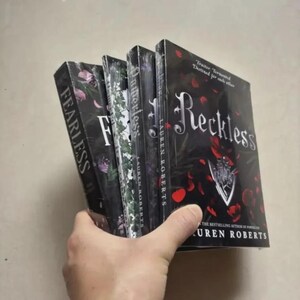 Pode incluir: Uma pilha de livros de Lauren Roberts, incluindo "Reckless" e "Fearless", segurada na m&atilde;o. As capas s&atilde;o predominantemente pretas com desenhos florais e de p&eacute;talas de rosa. O t&iacute;tulo "Reckless" est&aacute; em letras grandes e elegantes.