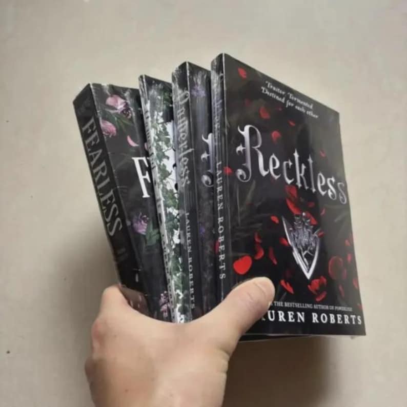 Pode incluir: Uma pilha de livros de Lauren Roberts, incluindo "Reckless" e "Fearless", segurada na m&atilde;o. As capas s&atilde;o predominantemente pretas com desenhos florais e de p&eacute;talas de rosa. O t&iacute;tulo "Reckless" est&aacute; em letras grandes e elegantes.
