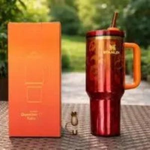 Puede incluir: Un vaso t&eacute;rmico rojo y dorado con asa y pajita, junto a una caja naranja. El vaso tiene la marca "Stanley". La caja tiene un dibujo del vaso.