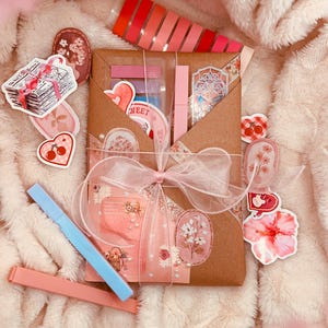 Puede incluir: Una colección de artículos de papelería, que incluye pegatinas, rotuladores y un paquete de regalo atado con una cinta rosa. Las pegatinas presentan diseños florales y de corazón, y los rotuladores son rosa pastel y azul.