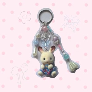 以下が含まれることがあります： チェック柄の服を着た小さなぬいぐるみウサギのキーホルダー。キーホルダーには、シルバーのリング、パステルカラーのビーズ、透明な蝶のチャーム、真珠色のドロップが含まれています。背景は白い水玉模様のピンク色です。
