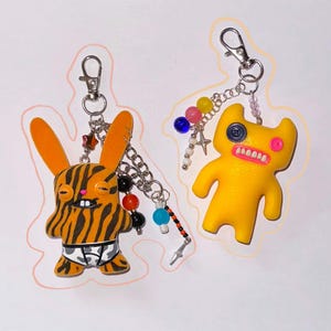 May include: Two handmade keyrings featuring quirky cartoon characters. One is a tiger-striped bunny wearing patterned underwear, the other a yellow monster with a button eye and pink mouth. Both have silver chain and bead accents.