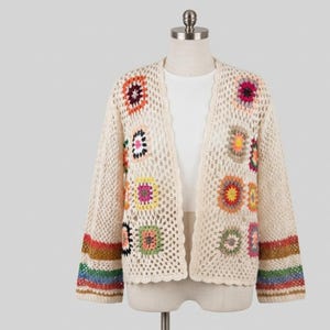 May include: A cream-colored open-front cardigan featuring a crochet construction with a net-like pattern. The garment is decorated with various multi-colored square motifs and finished with horizontal stripes in brown, green, and blue at the cuffs.