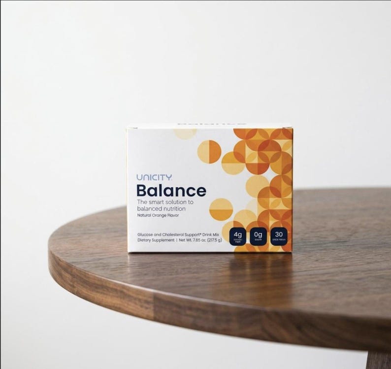 K&ouml;nnte beinhalten: Eine wei&szlig;e Schachtel mit Unicity Balance-Getr&auml;nkemix mit orangefarbenen und gelben geometrischen Designs. Auf der Schachtel steht "Balance" und "Die intelligente L&ouml;sung f&uuml;r ausgewogene Ern&auml;hrung". Die Schachtel steht auf einem Holztisch.