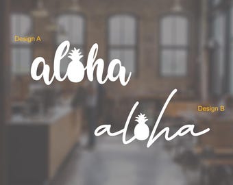 May include: Two white "aloha" designs with a pineapple graphic. Design A is above Design B. The background is blurred, showing a cafe setting with windows and a counter.