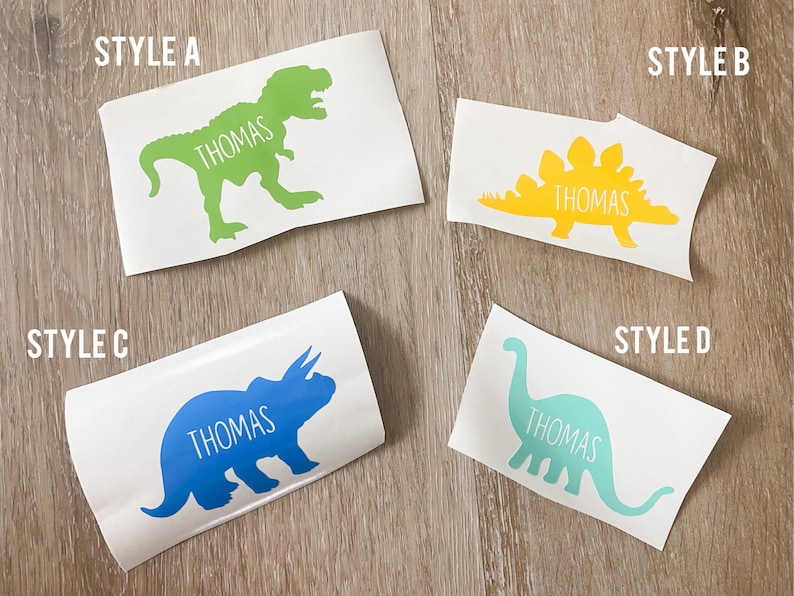 May include: Four different styles of dinosaur vinyl decals with the name "Thomas" on each. The decals are on a white background. Style A is a green T-Rex, Style B is a yellow Stegosaurus, Style C is a blue Triceratops, and Style D is a turquoise Brachiosaurus.