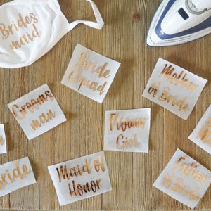 May include: A white face mask with the text Bridesmaid in metallic rose gold, accompanied by several iron-on transfer sheets featuring rose gold text including Bride Squad, Mother of Bride, Groomsman, Flower Girl, Best Man, Groom, Bride, Maid of Honor, and Father of Bride.