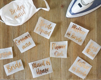 May include: A white face mask with the text Bridesmaid in metallic rose gold, accompanied by several iron-on transfer sheets featuring rose gold text including Bride Squad, Mother of Bride, Groomsman, Flower Girl, Best Man, Groom, Bride, Maid of Honor, and Father of Bride.
