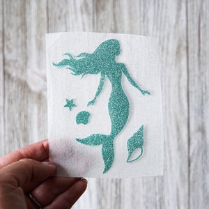 May include: A teal glitter mermaid silhouette with flowing hair, a starfish, a seashell, and a shell. The mermaid is on a white background. The image is held by a hand.