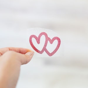 May include: A clear sheet of transfer paper with two pink glitter hearts on it. The hearts are overlapping and are in the shape of a double heart.