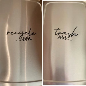 May include: Two stainless steel bins with labels. The left bin has a label that says "recycle" with a small leaf design. The right bin has a label that says "trash" with a small leaf design.