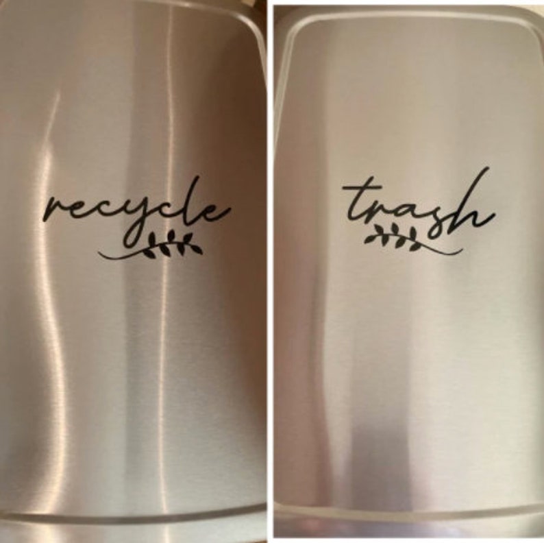 May include: Two stainless steel bins with labels. The left bin has a label that says "recycle" with a small leaf design. The right bin has a label that says "trash" with a small leaf design.