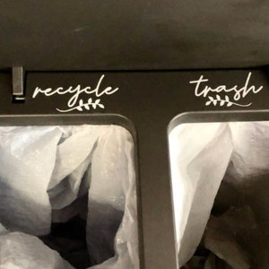 May include: A black bin with two compartments, labelled "recycle" and "trash" with a white leafy design. Each compartment has a white plastic bag inside.