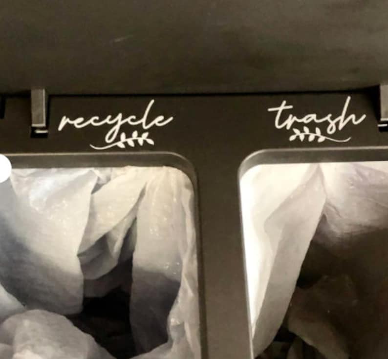 May include: A black bin with two compartments, labelled "recycle" and "trash" with a white leafy design. Each compartment has a white plastic bag inside.