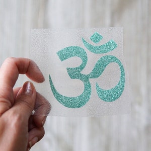 May include: A teal glitter Om symbol on a clear transfer sheet.