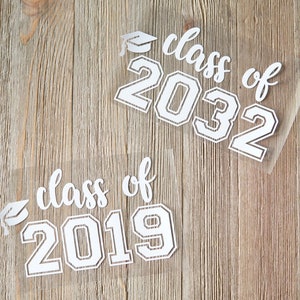 May include: Two white vinyl decals with the text "class of" and the year "2019" and "2032" in a bold font. The decals are on a wood background.