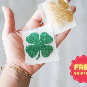 May include: Two glitter shamrock stickers, one green and one gold, on a white background. The stickers are on a person's hand. The text "FREE SHIPPING" is on a red background.