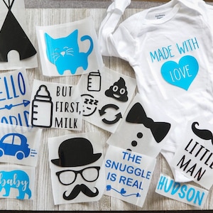 May include: A white long-sleeve baby bodysuit and a collection of vinyl heat transfer decals in black and blue. Designs include a teepee, cat, baby bottle, car, bear, mustache, bow tie, and text such as MADE WITH LOVE and THE SNUGGLE IS REAL.