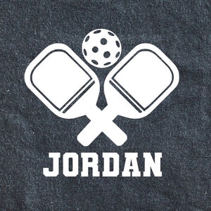 May include: White silhouette of two pickleball paddles crossed with a pickleball in the center. The name "JORDAN" is written below the paddles.