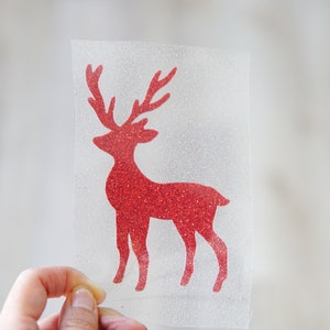 May include: A red glittery reindeer silhouette on a clear, textured background. The reindeer has large antlers and is standing. The image is held by a hand. This image is suitable for holiday crafts and decorations.