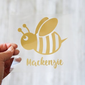 May include: Gold vinyl sticker with a bee design and the name Mackenzie. The bee is yellow with white stripes and has a black eye.