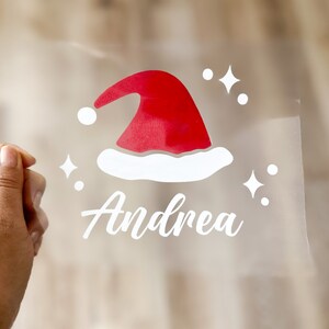 May include: White vinyl sticker with a red and white Santa hat and the name Andrea in white cursive lettering.