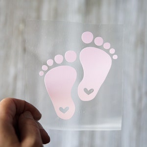May include: A clear vinyl sticker with two pink baby footprints with a small heart inside each footprint.
