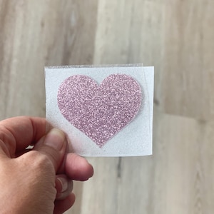 May include: A pink glitter heart-shaped iron-on transfer patch centered on a square piece of clear adhesive backing material. The heart features a dense, sparkling texture and is ready for application onto a fabric surface for decoration.