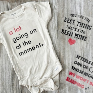 May include: A white onesie with black text that reads "a lot going on at the moment."  A clear vinyl sheet with black text that reads "You are the best thing that's ever been mine" with a red heart.  Another clear vinyl sheet with black text that reads "It feels like one of those nights we won't be sleeping."