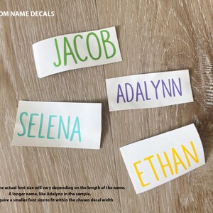 May include: Four custom name decals on a wooden surface. The names are Jacob, Selena, Adalynn, and Ethan. The decals are in different colours: green, blue, purple, and yellow.