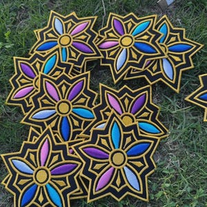 May include: A collection of embroidered flower patches. Each patch features a black background with a gold border and a central flower design. The flower petals are in shades of blue, purple, and pink, with a gold center. The patches are scattered on green grass.