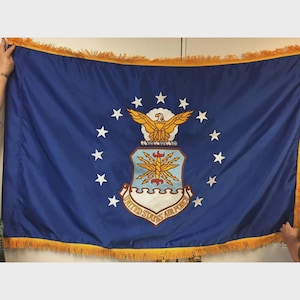 May include: A rectangular blue flag featuring the United States Air Force emblem in the center, surrounded by a circle of thirteen white stars. The emblem depicts a golden eagle above a shield with lightning bolts. The flag has gold fringe trim.