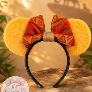 May include: A headband featuring two round, fuzzy, golden-yellow ears and a central fabric bow. The bow displays a repeating pattern of dark brown silhouettes of lions and geometric shapes on an orange background, secured by a jute-wrapped centre.