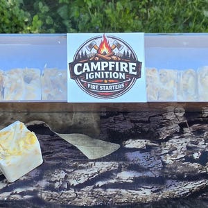 May include: A clear plastic box containing multiple fire starters, with one starter outside the box. The box has a label that reads "Campfire Ignition Fire Starters" with a graphic of a campfire. The fire starters appear to be made of a waxy material.