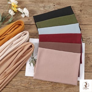 Pode incluir: Uma variedade de véus de tecido em várias cores, incluindo preto, verde-oliva, azul claro, bordô e marrom. Véus enrolados em tons de bege e dourado também são visíveis, dispostos sobre uma superfície de madeira.