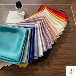 May include: A selection of folded satin scarves in a spectrum of colours, such as teal, blue, purple, pink, white, yellow, orange, and red. A perfume bottle and a card with text are also present.