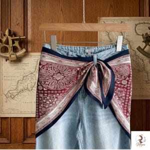 Könnte beinhalten: Hellblaue Jeans aus Denim mit einem gebundenen Seidentuch in Burgunderrot und Weiß mit Paisleymuster, das über die vordere Taille drapiert ist. Das Tuch hat einen dunkelblauen Rand und ist mittig geknotet, was einen Lagenlook erzeugt.