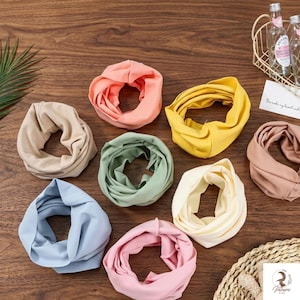 May include: A collection of eight circular fabric headbands in various solid colours including peach, yellow, beige, sage green, light blue, cream, pink, and dusty rose, arranged on a wooden surface with a wire basket and a small white card.