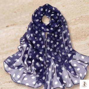 May include: A sheer navy blue scarf featuring a repeating pattern of white polka dots. The lightweight fabric is draped to show its translucent quality and soft folds. A small white rectangular tag is visible near the center of the scarf.