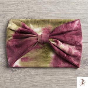 Könnte beinhalten: Ein breites Stoffstirnband mit einem Batikmuster in den Farben tiefes Burgunderrot und gedämpftes Olivgrün. Das Stirnband ist in der Mitte mit einem Stoffknotendetail gerafft, was eine weiche, gefaltete Optik auf der gesamten Oberfläche erzeugt.
