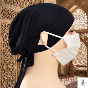 May include: A solid black head covering featuring a tie-back closure at the nape of the neck. The fabric is gathered at the back to create a soft, draped silhouette. A white disposable pleated face mask is worn over the ears with white elastic loops.