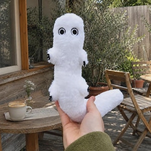 Pode incluir: Um brinquedo de pelúcia branco com um corpo longo e cilíndrico, uma cauda grossa e dois braços pequenos. O brinquedo apresenta dois olhos grandes e circulares bordados em preto e branco e pequenas garras pretas nas mãos e pés de tecido macio.