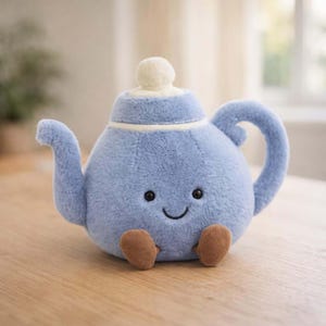 Puede incluir: Una tetera de peluche azul claro con una cara sonriente, ojos negros y pequeños pies marrones. La tetera tiene una tapa blanca y un asa curva. Un juguete para niños.